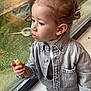 child, toddler, denim_jacket, snack, bread, crumbs, window, glass_reflection, hair_bun, side_profile, looking_out, indoor, greenery_outside, sitting, hand, shoe, pants, face, portrait, casual_clothing