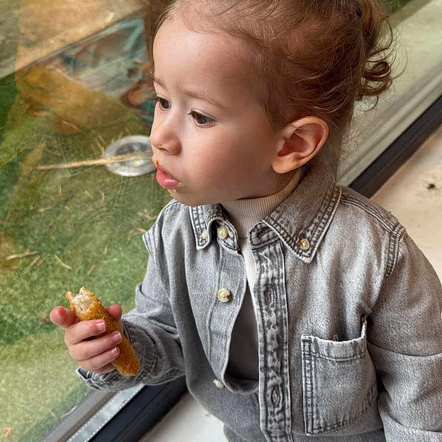 Shay a rejoint le concours — aidez-le/la à gagner de superbes lots ! bread, casual_clothing, child, crumbs, denim_jacket, face, glass_reflection, greenery_outside, hair_bun, hand, indoor, looking_out, pants, portrait, shoe, side_profile, sitting, snack, toddler, window