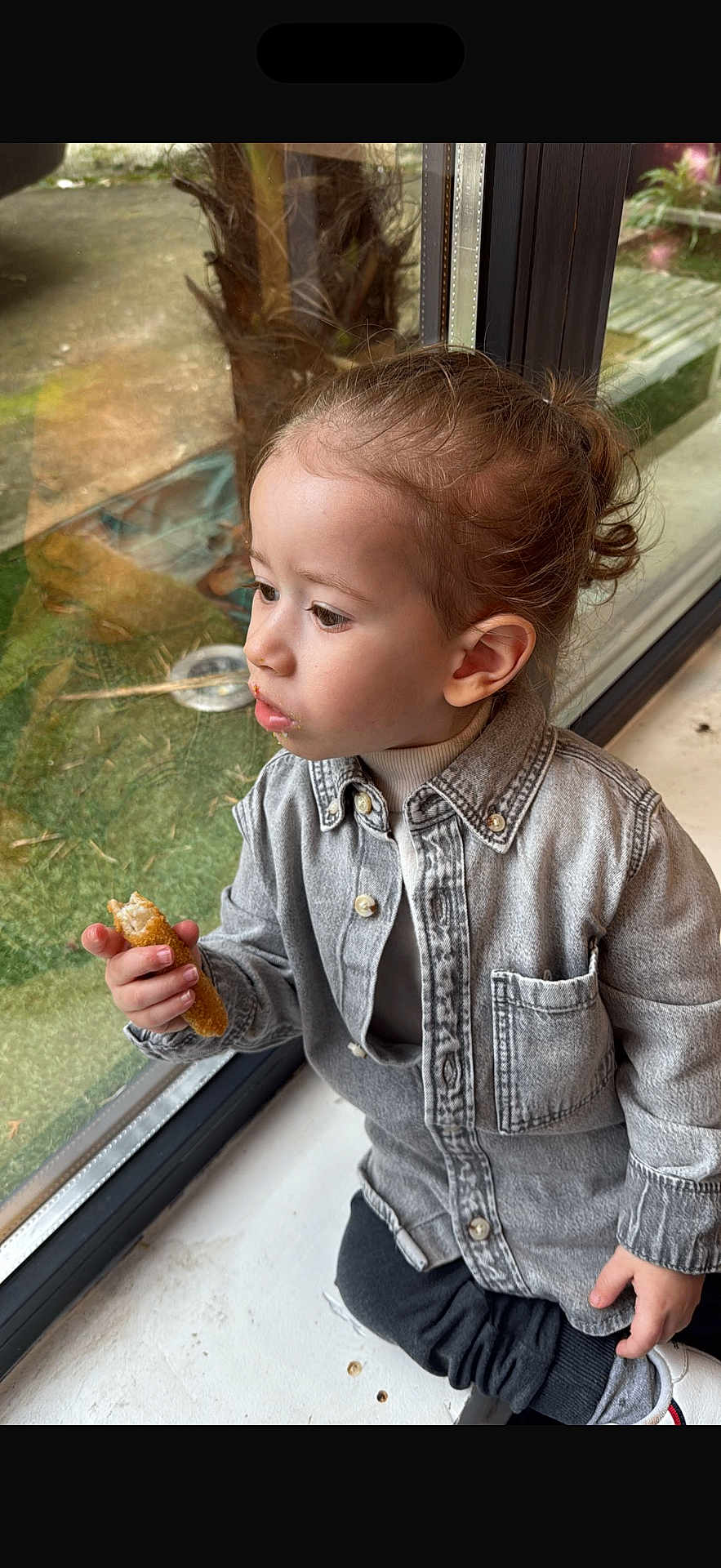 Shay a rejoint le concours — aidez-le/la à gagner de superbes lots ! child, toddler, denim_jacket, snack, bread, crumbs, window, glass_reflection, hair_bun, side_profile, looking_out, indoor, greenery_outside, sitting, hand, shoe, pants, face, portrait, casual_clothing