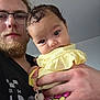baby, infant, child, adult, man, beard, glasses, pacifier, hand, holding, yellow_clothing, selfie, portrait, closeup, indoor, wall, hair, eyes, face, curious