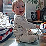 baby, child, pajamas, smiling, sitting, toy, play_mat, indoor, sunlight, plant, furniture, happy, face, floor, container, stuffed_toy, blanket, window, home, decor
