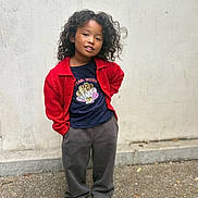 Hilona is registered to the contest to win money with this photo: child, portrait, standing, outdoors, sidewalk, wall, red_cardigan, dark_shirt, gray_pants, white_shoes, curly_hair, smiling, fashion, casual_clothing, full_body, young_person, street_style, urban, daylight, pavement