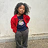 child, portrait, standing, outdoors, sidewalk, wall, red_cardigan, dark_shirt, gray_pants, white_shoes, curly_hair, smiling, fashion, casual_clothing, full_body, young_person, street_style, urban, daylight, pavement