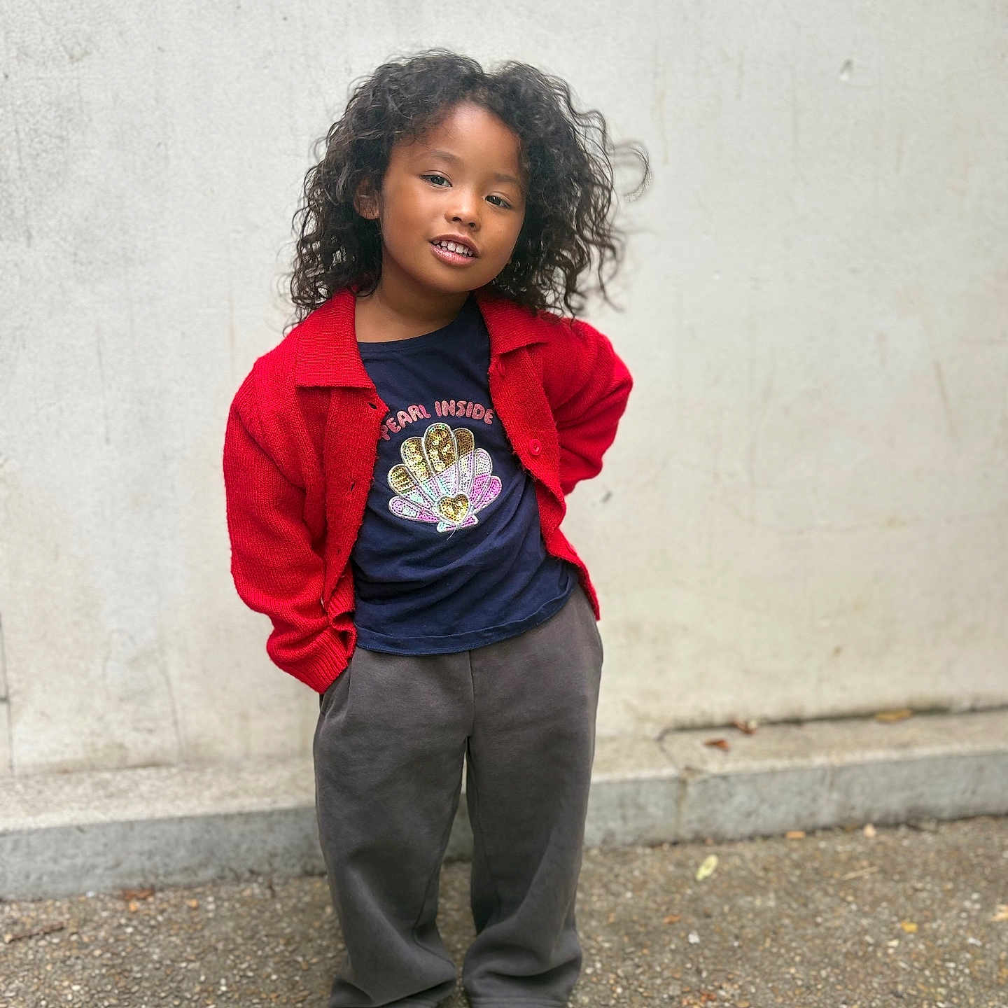 Hilona is registered to the contest to win money with this photo: child, portrait, standing, outdoors, sidewalk, wall, red_cardigan, dark_shirt, gray_pants, white_shoes, curly_hair, smiling, fashion, casual_clothing, full_body, young_person, street_style, urban, daylight, pavement