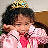 chair, child, clothing, crown, curly_hair, cute, girl, hand_on_face, happy, headwear, indoor, jewelry, pink_jacket, pink_pajamas, playful, portrait, smile, teeth, tiara, young_child