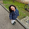 bicycle_wheel, child, coat, concrete, curb, curly_hair, grass, jacket, kid, moss, outdoor, pavement, playful, portrait, shoes, smiling, sneakers, toddler, tree, wall