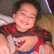 Marvin is registered to the contest to win money with this photo: toddler, child, smile, remote_control, blanket, curly_hair, indoor, happy, person, hand, red_clothing, blue_blanket, yellow_blanket, candid, closeup, face, dim_light, holding, cozy, infant