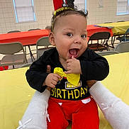 Jeremiah is registered to the contest to win money with this photo: baby, birthday, crown, smile, happy, child, red_pants, yellow_bow, black_shirt, party, indoor, table, chair, celebration, person, hands, festive, cute, first_birthday, infant