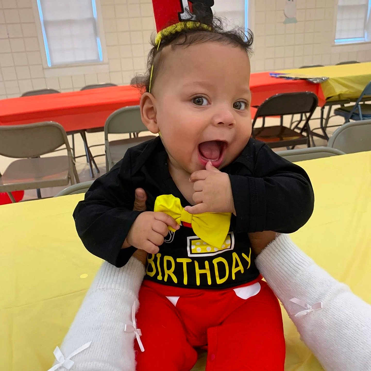 Jeremiah is registered to the contest to win money with this photo: baby, birthday, crown, smile, happy, child, red_pants, yellow_bow, black_shirt, party, indoor, table, chair, celebration, person, hands, festive, cute, first_birthday, infant