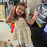 Athena Joseph is registered to the contest to win money with this photo: child, girl, smiling, peace_sign, dress, floral_pattern, indoors, toys, colorful_blocks, sandals, floor, furniture, storage_boxes, happy, playful, person, portrait, curly_hair, casual, home