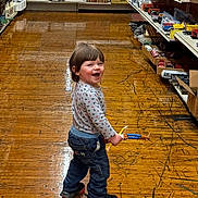 Vincent is registered to the contest to win money with this photo: aisle, casual_wear, child, clothing, floor_reflection, happy, indoor, jeans, long_sleeve_shirt, person, playful, retail, shelves, smiling, standing, store, toddler, toy, wooden_floor, young_child