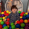 ball_pit, balls, bright_colors, casual_clothing, child, colorful, face, fun, happy, indoor, leisure, person, play, playpen, seated, smiling, striped_shirt, toddler, toy, young_child