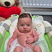 George is registered to the contest to win money with this photo: baby, bathtub, bubbles, teddy_bear, red_heart, water, foam, infant, bath_time, toy, child, indoor, carpet, radiator, skin, head, face, expression, sitting, cute