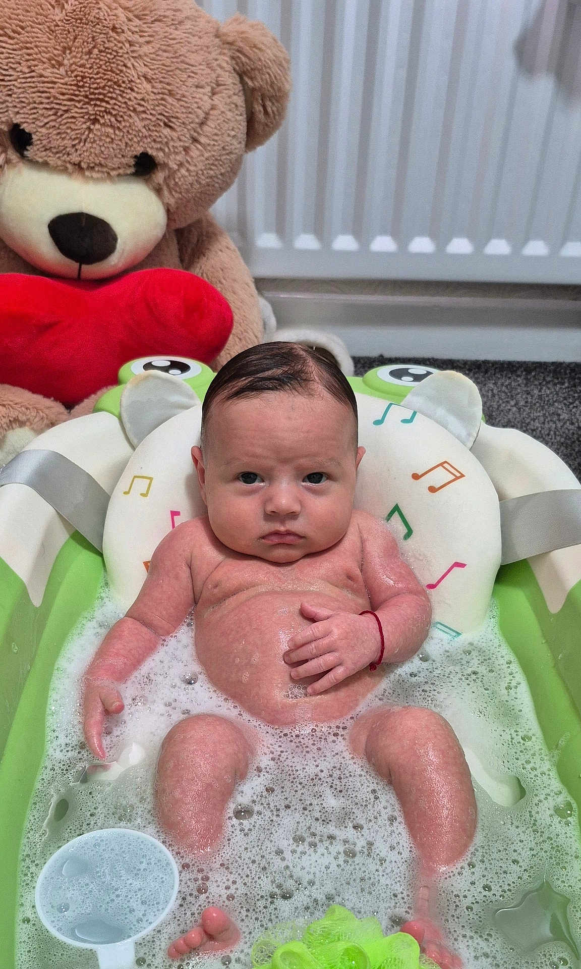 George is registered to the contest to win money with this photo: baby, bathtub, bubbles, teddy_bear, red_heart, water, foam, infant, bath_time, toy, child, indoor, carpet, radiator, skin, head, face, expression, sitting, cute