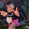 Mariana is registered to the contest to win money with this photo: child, girl, peace_sign, hello_kitty, sweatshirt, pink_shorts, folding_chair, outdoor, grass, s'more, snack, camouflage, smile, person, night, relaxed, hand_gesture, playful, young, casual