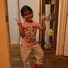 child, girl, indoor, peace_sign, smile, pink_shirt, red_shoes, casual_clothing, wooden_floor, cabinet, glass, decor, happy, posing, person, home, flooring, toy, cute, young