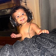 Alexia is registered to the contest to win money with this photo: baby, child, headphones, smiling, indoor, portrait, face, person, infant, happy, curly_hair, blanket, fuzzy, home, furniture, fireplace, curtain, sitting, music, cute