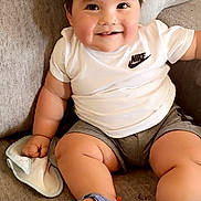 Jeremiah joined the competition — help win amazing prizes! baby, child, smiling, sitting, white_shirt, nike_logo, gray_shorts, blue_sandals, couch, soft_furniture, indoors, cute, happy, chubby, infant, holding_cloth, short_hair, skin, casual_clothing, portrait
