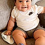 baby, child, smiling, sitting, white_shirt, nike_logo, gray_shorts, blue_sandals, couch, soft_furniture, indoors, cute, happy, chubby, infant, holding_cloth, short_hair, skin, casual_clothing, portrait