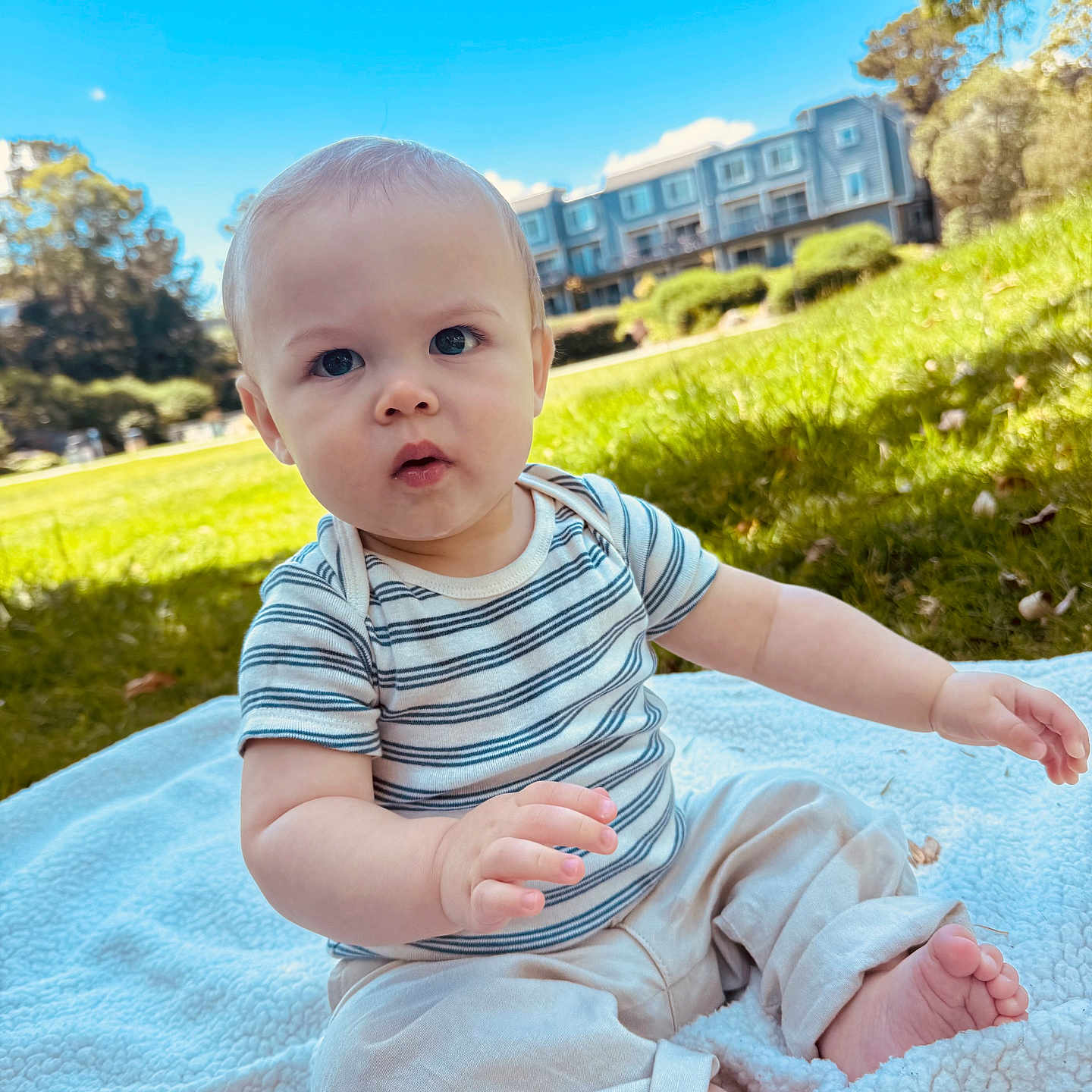 Christian is registered to the contest to win money with this photo: baby, child, infant, outdoor, grass, blue_sky, tree, blanket, striped_shirt, pants, barefoot, greenery, daylight, building, nature, curious, sitting, cute, portrait, person