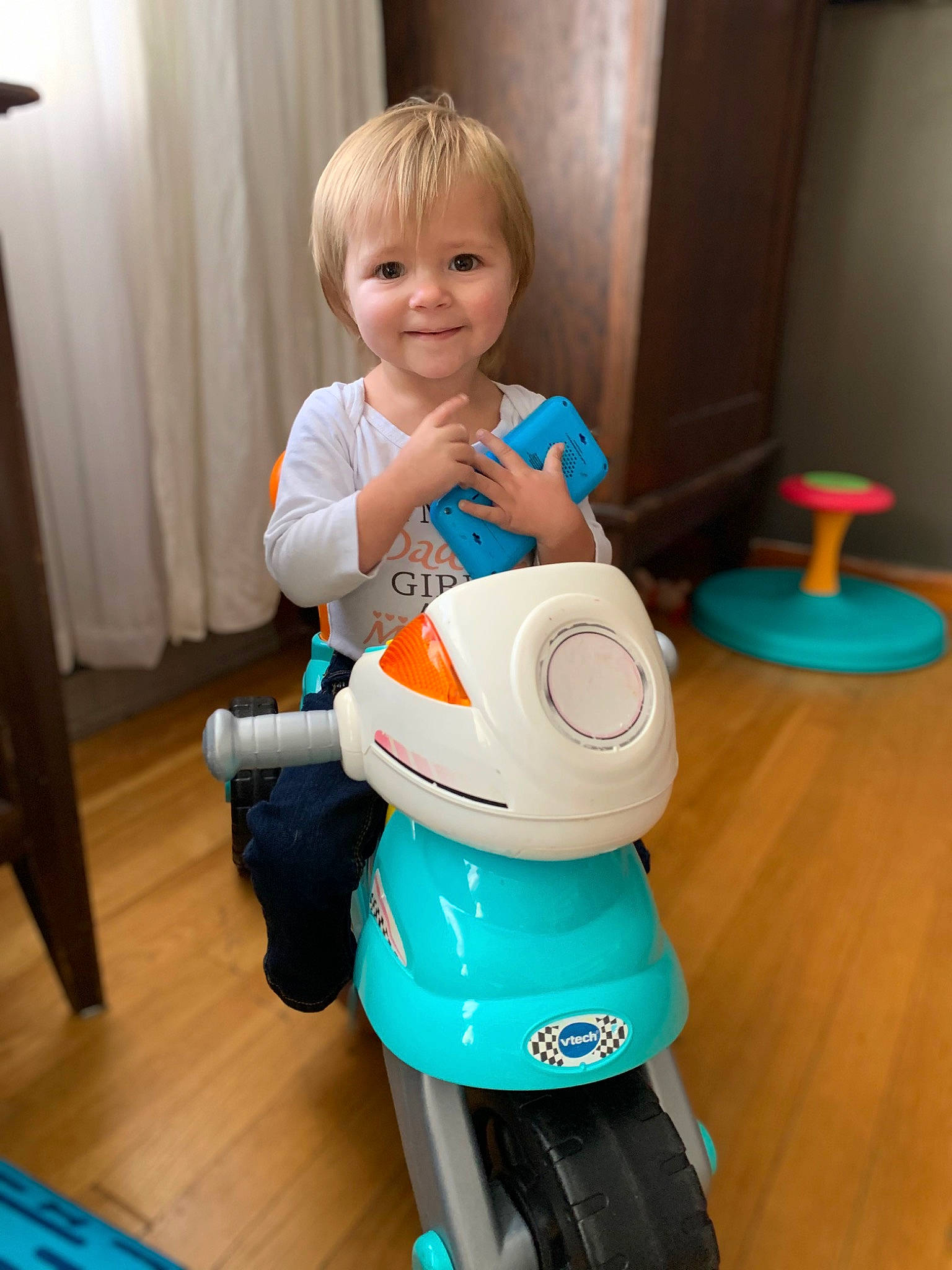 Callie is registered to the contest to win money with this photo: baby_products, baby_toddler_clothing, baby_toys, blue, child, cleanliness, curtain, electric_blue, fun, happy, joy, person, plastic, play, product, riding_toy, smile, standing, t_shirt, tire