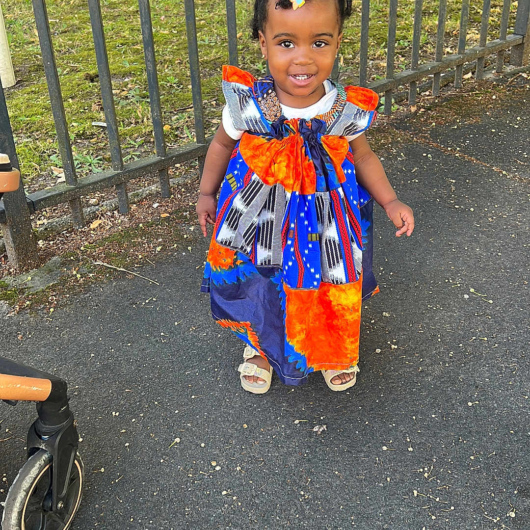 Parfaite Grâce Olga participe au concours pour gagner de l'argent avec cette photo : child, colorful_clothing, daylight, dress, fence, grass, hair_clip, happy, nature, outdoor, person, portrait, sandals, sidewalk, smiling, standing, stroller, toddler, tree, young_child
