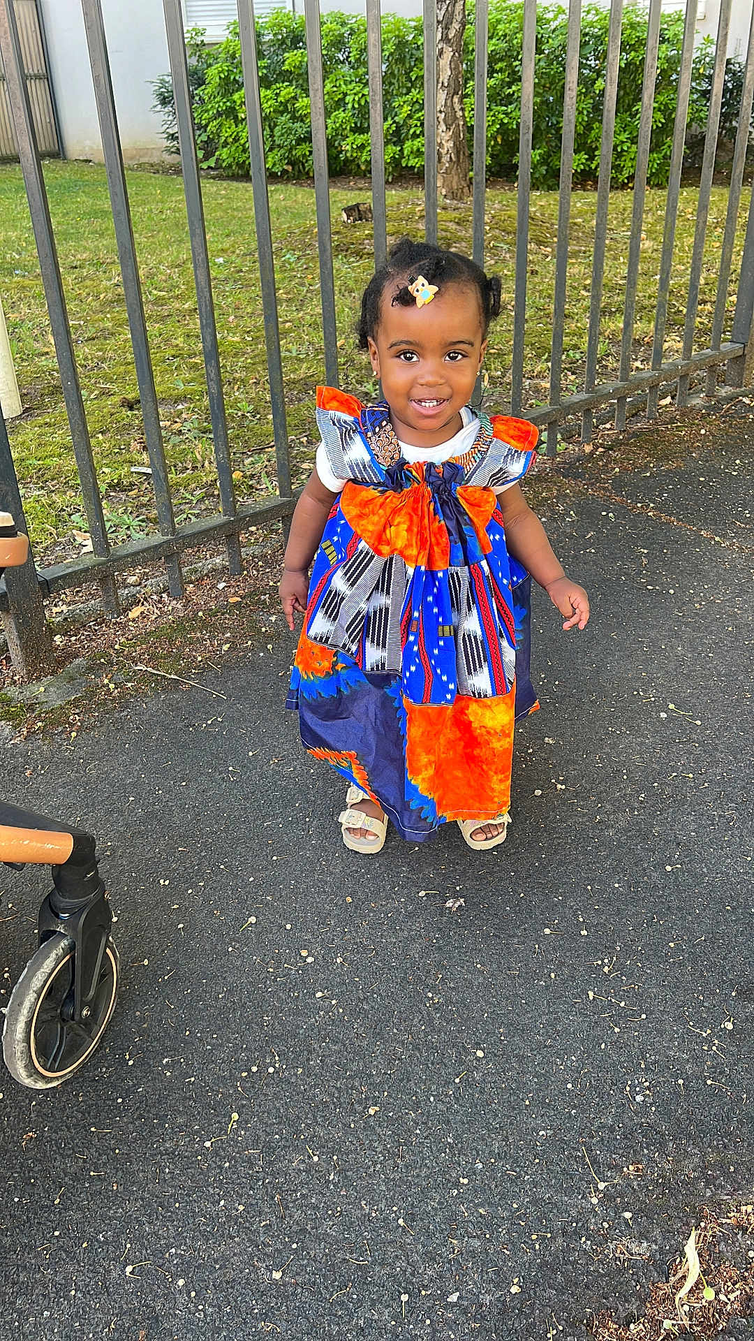 Parfaite Grâce Olga participe au concours pour gagner de l'argent avec cette photo : child, colorful_clothing, daylight, dress, fence, grass, hair_clip, happy, nature, outdoor, person, portrait, sandals, sidewalk, smiling, standing, stroller, toddler, tree, young_child