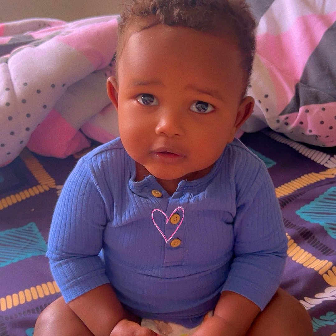 Sadam participe au concours pour gagner de l'argent avec cette photo : toddler, child, baby, bed, blanket, pillow, indoors, blue_clothing, diaper, curly_hair, skin, face, hands, sitting, cute, expression, portrait, colorful, soft_lighting, comfort