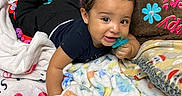 Jahkai is registered to the contest to win money with this photo: baby, child, infant, smiling, pacifier, lying_down, blanket, bed, bedroom, portrait, curly_hair, dark_clothing, colorful, cozy, cushion, textile, indoors, happy, playful, close_up
