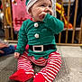 Maël participe au concours pour gagner de l'argent avec cette photo : baby, child, elf_costume, striped_leggings, red_socks, green_top, hat, holiday, festive, carpet, indoor, playful, cute, smiling, young_child, person, seated, clothing, face, hand
