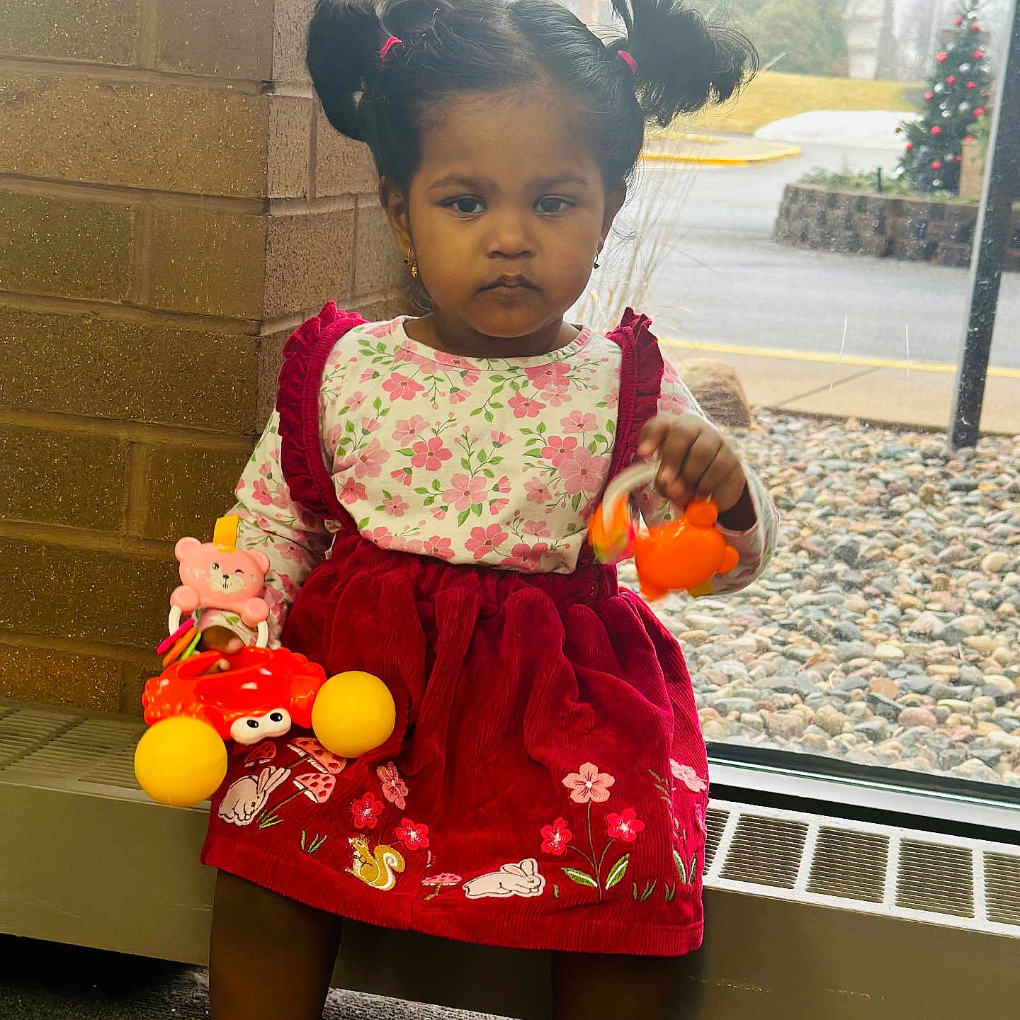 Moksha is registered to the contest to win money with this photo: brick_wall, carpet, child, decorations, dress, floral_pattern, girl, indoor, outside, person, pigtails, radiator, red_dress, rocks, serious_expression, shoes, toddler, toys, window, winter