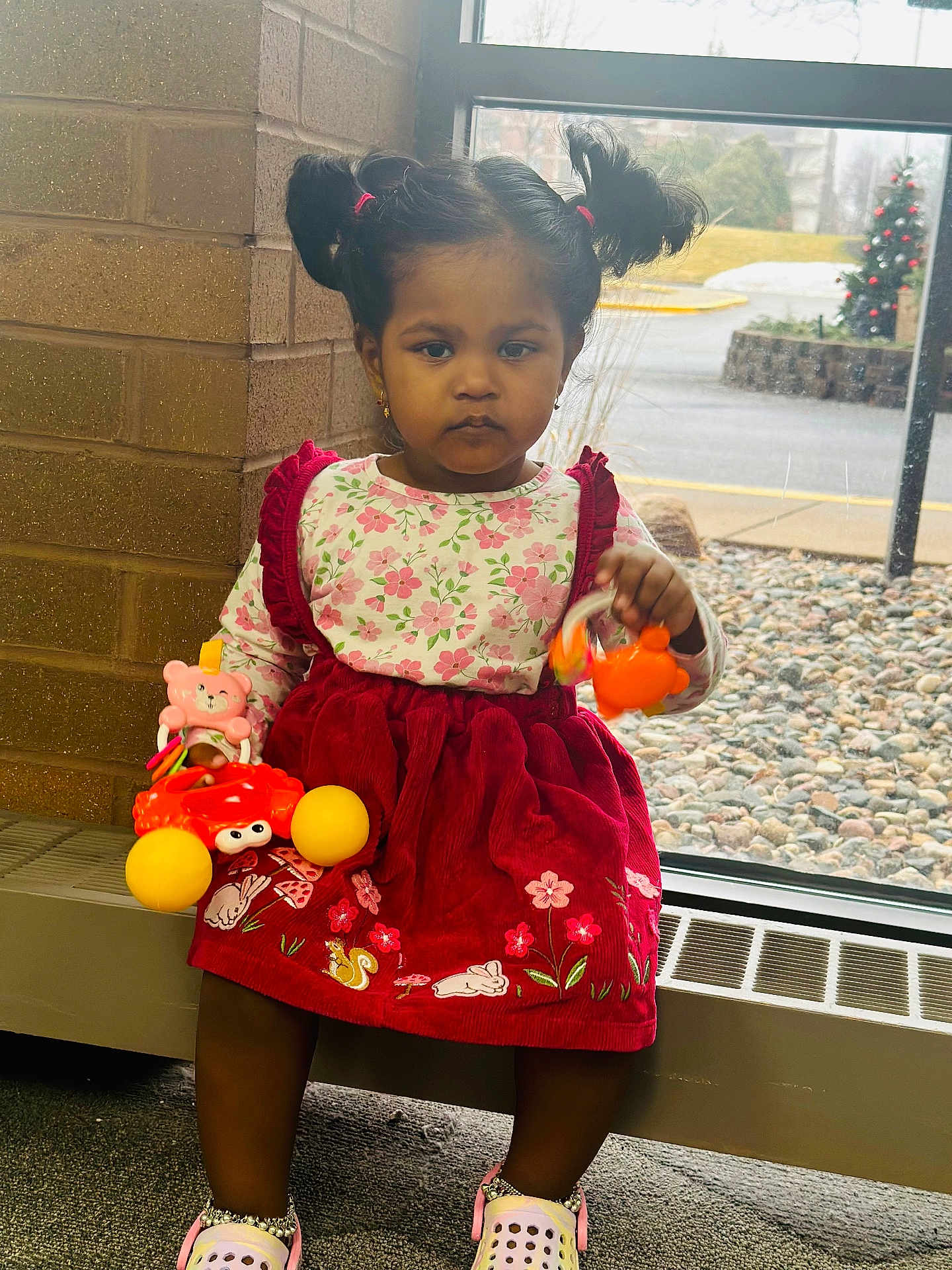 Moksha is registered to the contest to win money with this photo: toddler, child, girl, indoor, window, toys, pigtails, dress, red_dress, floral_pattern, serious_expression, shoes, carpet, radiator, brick_wall, winter, outside, rocks, decorations, person