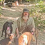 man, beard, glasses, cap, dog, dogs, outdoor, forest, dirt_path, sunlight, trees, leaves, pets, animal, walking, nature, canine, friendship, companion, casual