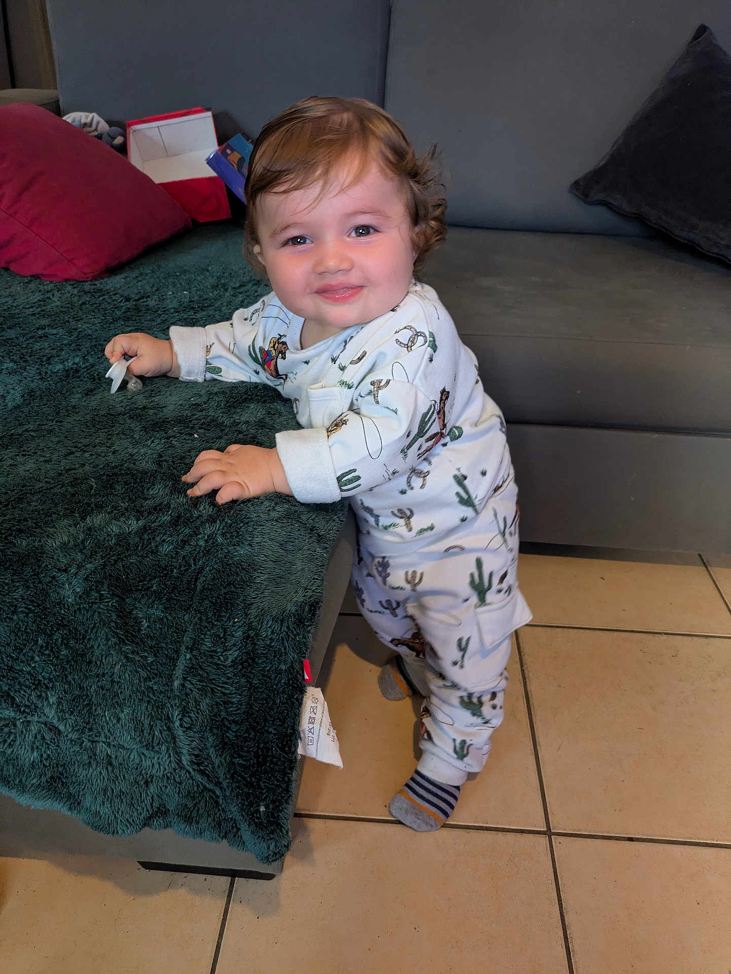 Gabriel participe au concours pour gagner de l'argent avec cette photo : toddler, child, smile, curly_hair, onesie, cactus_pattern, socks, blanket, couch, indoor, tile_floor, furniture, playful, standing, baby, portrait, happy, home, cozy, cute