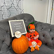 Zackary is registered to the contest to win money with this photo: baby, pumpkin, baby_costume, orange_clothing, couch, gray_furniture, sign, celebration, smiling, indoor, holiday, halloween, child, cute, plush, sitting, headwear, decor, three_months, festive