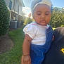 Madison is registered to the contest to win money with this photo: baby, child, outdoor, daylight, headband, flower, denim_overalls, grass, bush, house, cloud, sky, person, hand, holding, serious_face, short_sleeves, white_shirt, nature, sunlight