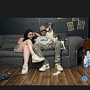 Milo joined the competition — help win amazing prizes! couch, dog, person, tattoo, sneakers, floor, poster, cap, camouflage_pants, living_room, wooden_floor, casual, indoor, relaxed, seated, pet, black_wall, sock, furniture, people