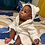 baby, child, infant, robe, white_robe, hood, bed, bedspread, blue_spoon, tissue, foot, toes, curious, wide_eyes, indoor, soft_texture, plush, cute, sitting, person