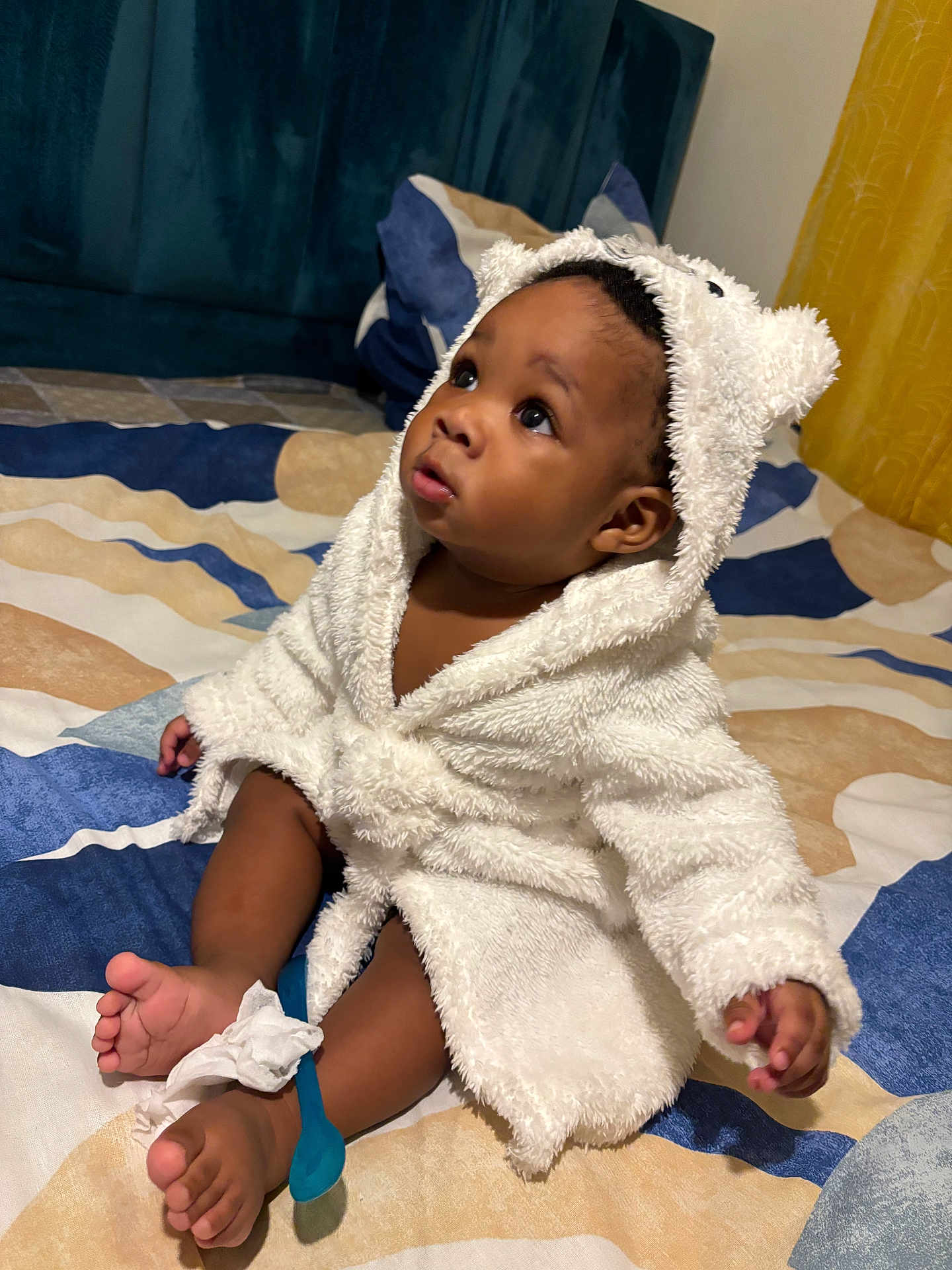 Sidiki participe au concours pour gagner de l'argent avec cette photo : baby, child, infant, robe, white_robe, hood, bed, bedspread, blue_spoon, tissue, foot, toes, curious, wide_eyes, indoor, soft_texture, plush, cute, sitting, person