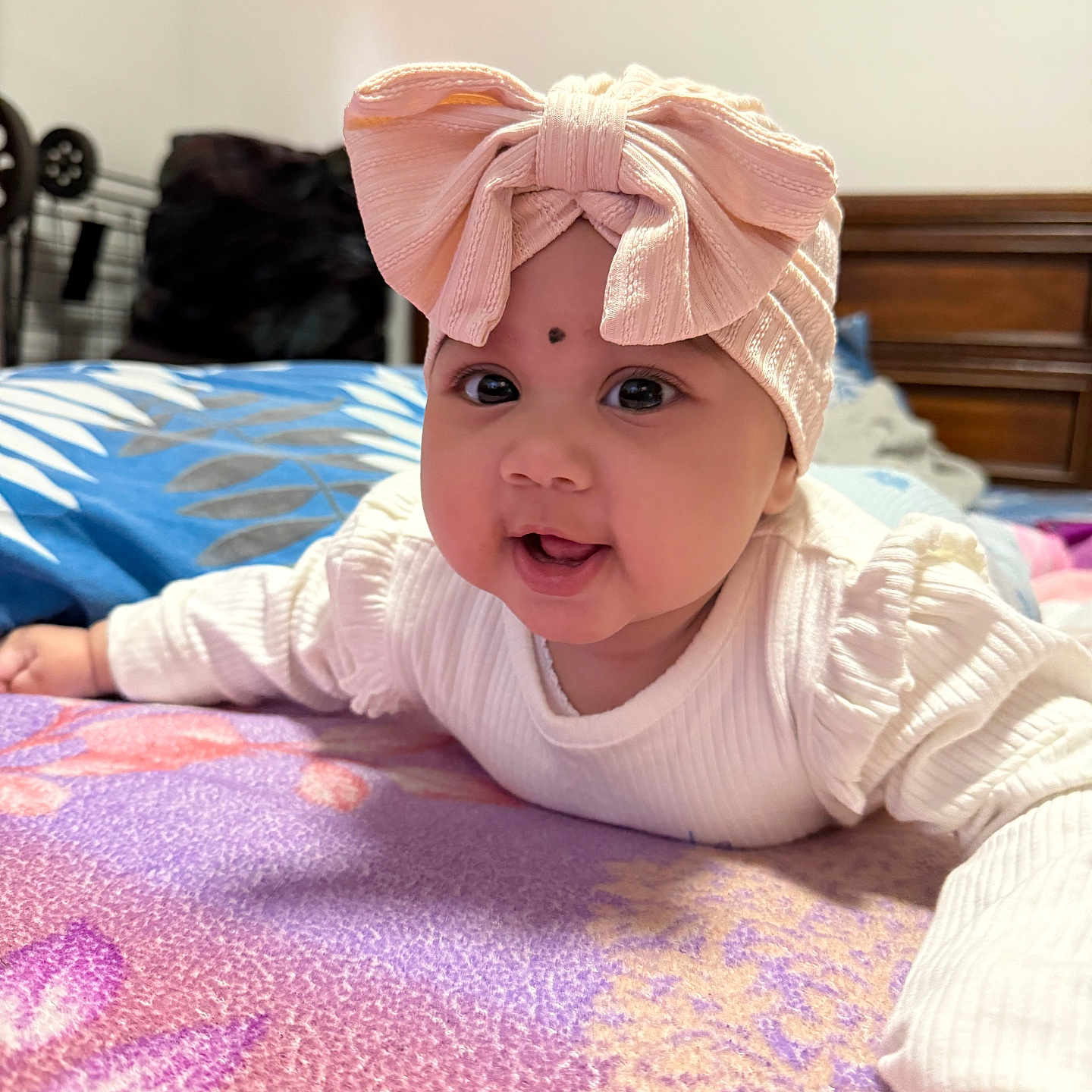 Sarah joined the competition — help win amazing prizes! baby, child, headband, pink_bow, white_clothing, blanket, indoor, bedroom, face, cute, smiling, toddler, lying_down, bright_eyes, curious, soft_lighting, closeup, portrait, colorful_blanket, cozy