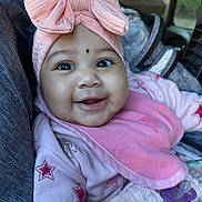 Sarah is registered to the contest to win money with this photo: baby, smiling, pink_bow, pacifier, car_seat, clothing, bib, star_pattern, infant, face, child, cute, happy, indoors, seat, headwear, portrait, closeup, young_child, cozy