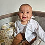 Tiago a rejoint le concours — aidez-le/la à gagner de superbes lots ! baby, child, smile, happy, infant, clothing, white_shirt, brown_pants, playpen, patterned_bedding, barefoot, indoors, cute, portrait, sitting, person, young_child, face, eyes, joy