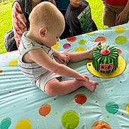Kadence is registered to the contest to win money with this photo: baby, child, people, cake, birthday_cake, tablecloth, balloon_pattern, outdoor, grass, smiling, curious, sitting, clothing, person, head, face, young_child, celebration, candle, colorful