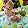baby, child, people, cake, birthday_cake, tablecloth, balloon_pattern, outdoor, grass, smiling, curious, sitting, clothing, person, head, face, young_child, celebration, candle, colorful