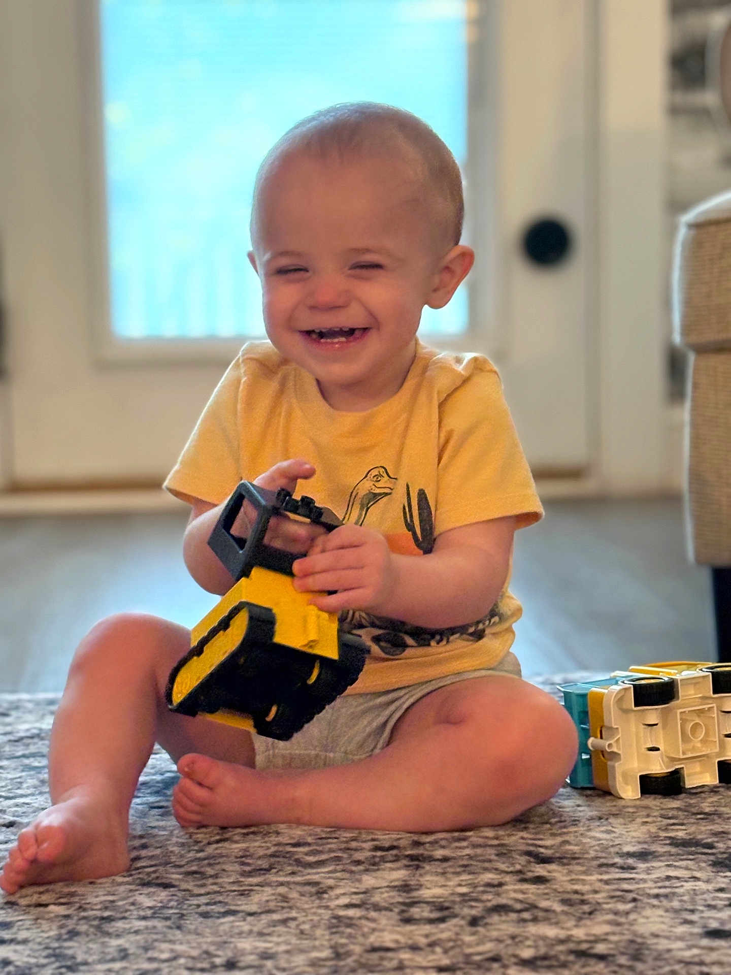 Kadence is registered to the contest to win money with this photo: toddler, child, smiling, toy, yellow_toy, truck, carpet, indoor, sitting, happy, shorts, tshirt, barefoot, playtime, home, daylight, candid, cute, young_child, floor