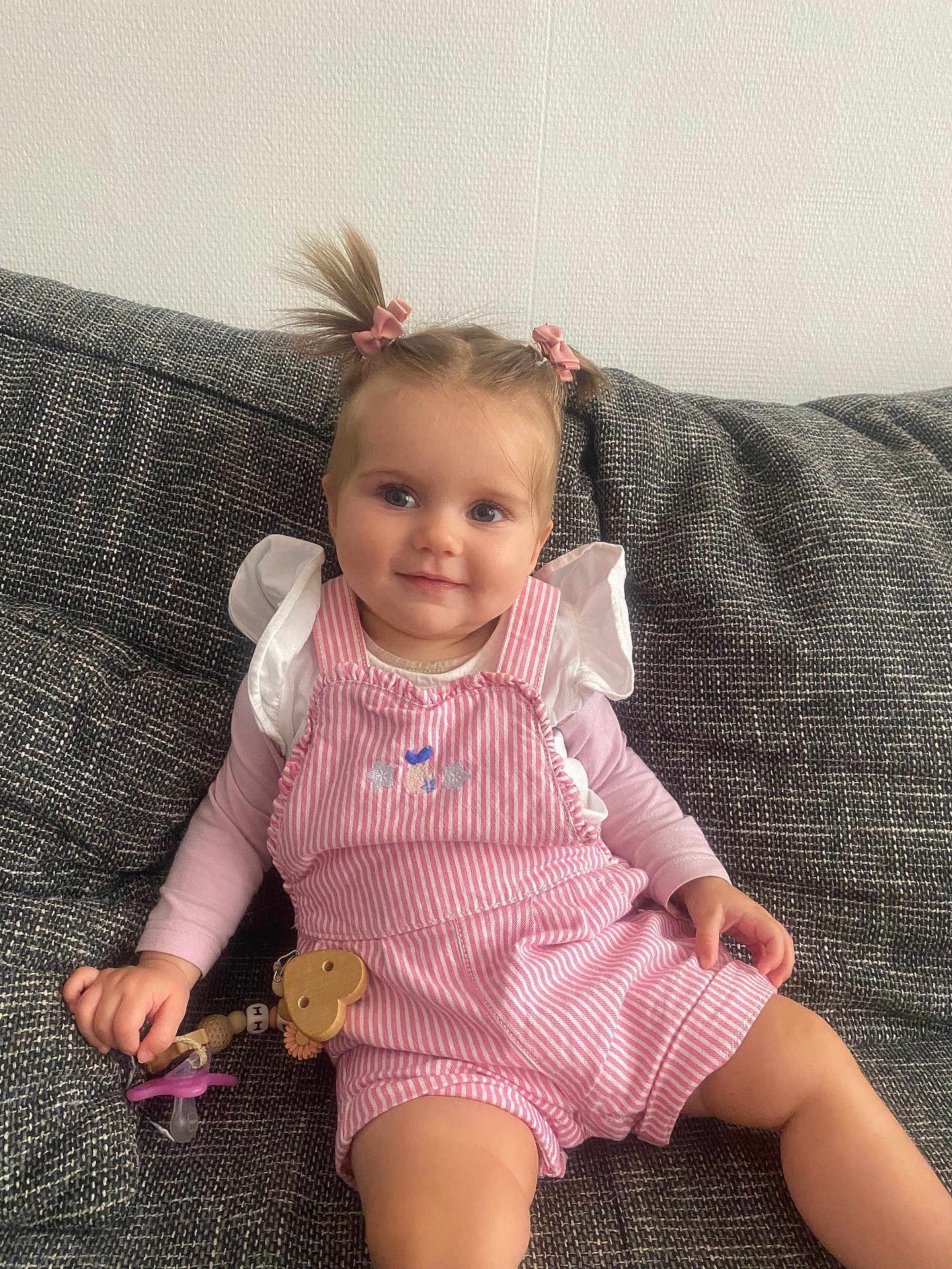 Iris a rejoint le concours — aidez-le/la à gagner de superbes lots ! toddler, child, pigtails, pink_bows, striped_romper, long_sleeve_shirt, pacifier, wooden_heart, couch, textured_fabric, smile, face, hair, sitting, indoors, cute, baby, hand, furniture, portrait