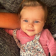 Iris participe au concours pour gagner de l'argent avec cette photo : baby, child, smiling, blue_eyes, ponytail, pink_shirt, floral_overall, cushion, blanket, cozy, indoor, portrait, cute, infant, happy, closeup, person, soft_lighting, relaxed, home