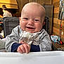 baby, smiling, high_chair, indoor, wooden_walls, kitchen, person, infant, clothing, hand, face, happy, young_child, background_person, cabinet, refrigerator, blanket, cozy, zipper, home