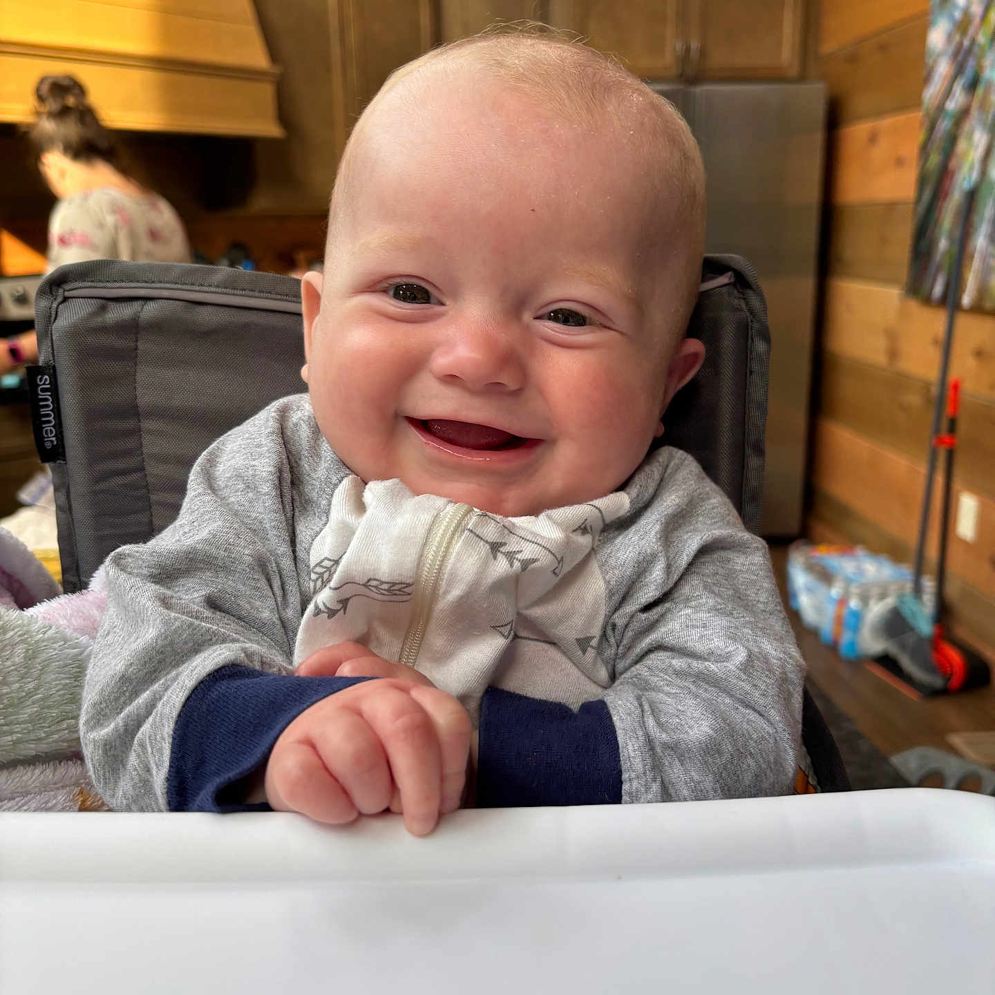 Styles is registered to the contest to win money with this photo: baby, background_person, blanket, cabinet, clothing, cozy, face, hand, happy, high_chair, home, indoor, infant, kitchen, person, refrigerator, smiling, wooden_walls, young_child, zipper