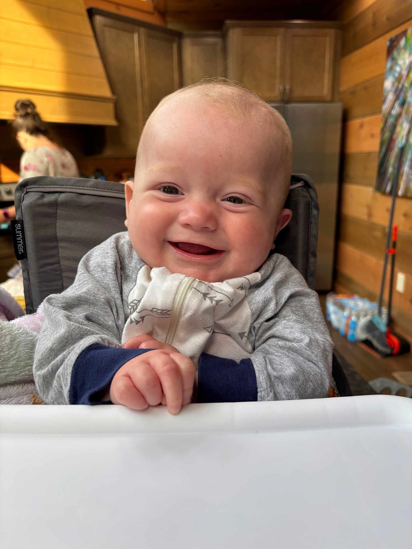 Styles is registered to the contest to win money with this photo: baby, smiling, high_chair, indoor, wooden_walls, kitchen, person, infant, clothing, hand, face, happy, young_child, background_person, cabinet, refrigerator, blanket, cozy, zipper, home